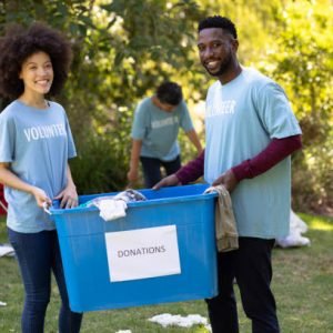 Mixed race couple spending time outside with their family, presenting a donations box with clothes in it, looking at the camera and smiling, with their family in the background, all wearing blue volunteers t shirts, on a sunny day